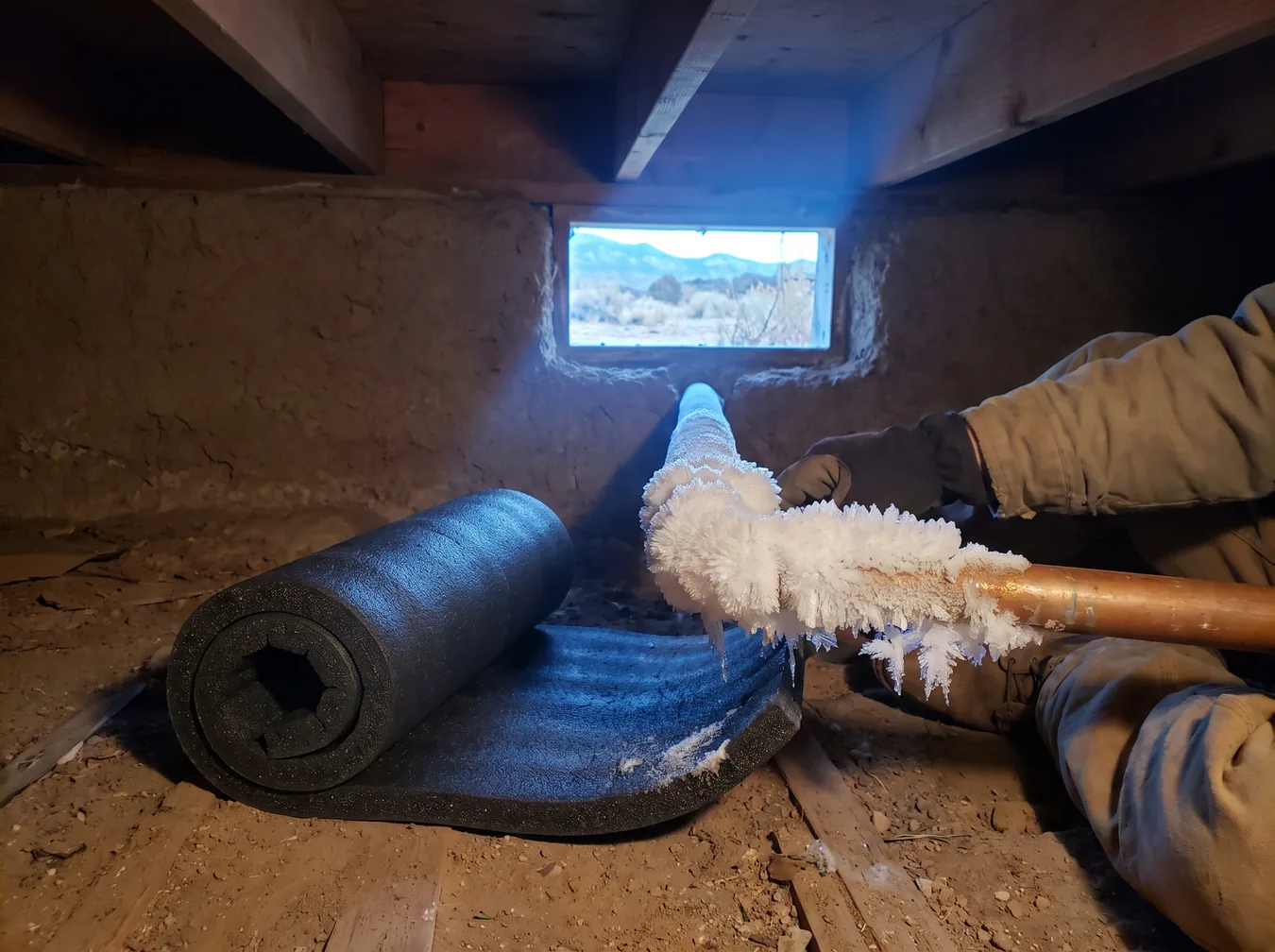 Frost-covered copper water pipe in a crawlspace next to foam pipe insulation